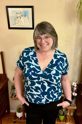 Sarah stands smiling at the camera with her hands in her pockets of her ready to wear black trousers.  She is wearing a Turquoise short-sleeved Zakopane Top from Itch to Stitch