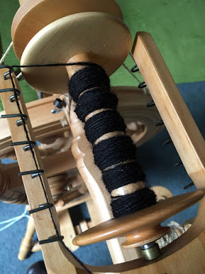 View from above of a bobbin on a castle-style spinning wheel, all in light, natural wood. Six neat bumps of tightly-spun black yarn are wound on the core of the bobbin, and the yarn is hooked around the furthest hook of the flyer from the photographer, running along all the other hooks and out of frame.