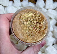 A white hand holding a cylindrical clear tub that is open showing a thick paste like golden brown sugar scrub with golden sugar running throughout with a gold glitter top on a bright background