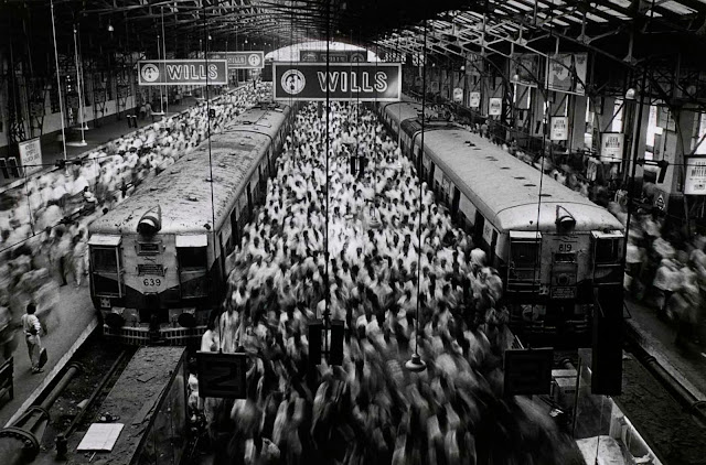 Foto em preto e branco, mostra uma estação de trem, com dois trens, e a imagem borrada de várias pessoas andando por lá