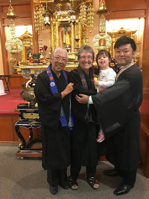 Three Buddhist ministers in robes holding a smiling child while standing in front of a Buddhist altar