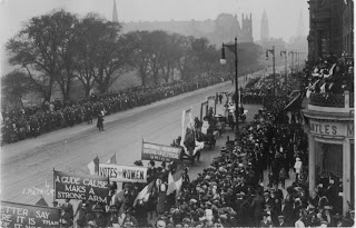 A black and white photograph of a procession of people carrying banners saying, for example, Votes for Women, they are marching diagonally from the top right of the frame to the bottom left and the Edinburgh skyline is visible in the background. 