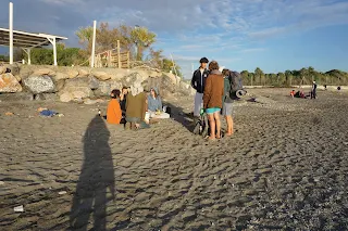 About 10 people sitting and standing in the sand on a beach and talking