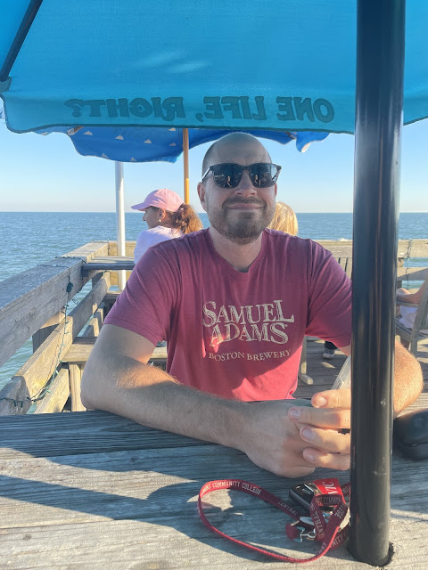 A picture of dad with his sunglasses on smiling at the camera at the picnic table of the restaurant we ate at on that first night. The ocean is behind him.