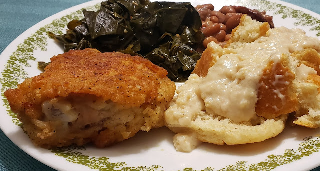 White plate with green flowers, fried chicken, collard greens, pinto beans and gravy and biscuits