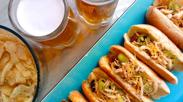 Game Day Jalapeno Cheddar Bacon Dogs on a serving tray next to a bowl of chips and two glasses of beer