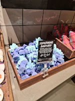 A photo of a light brown square box containing a bunch of green and blue screaming banshee shaped bath bombs next to a black rectangular card that says screaming banshee bath bomb in white font on a light brown rectangular table on a bright background