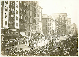 A black and white photograph of a parade moving through a crowded street.