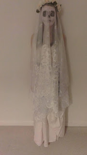 Me wearing my homemade bride of the dead Halloween costume, stood ghostly against white wall, with net curtain veil over face, secured with flower crown. Draped white bed sheets over black poofy dress to create off-white wedding dress look.