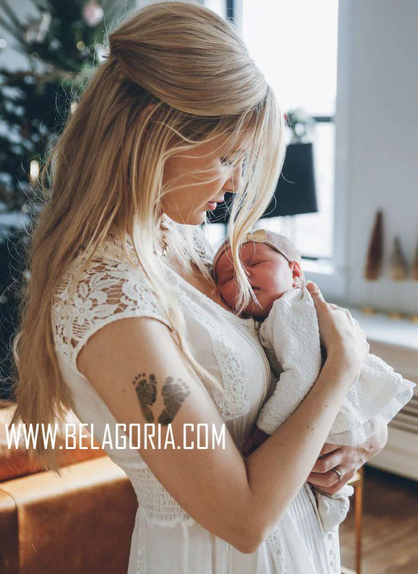 vemos a una madre de cabello rubio y vestido blanco acunando en brazos a su bebe, lleva un tatuaje con las huellas del bebe en el brazo