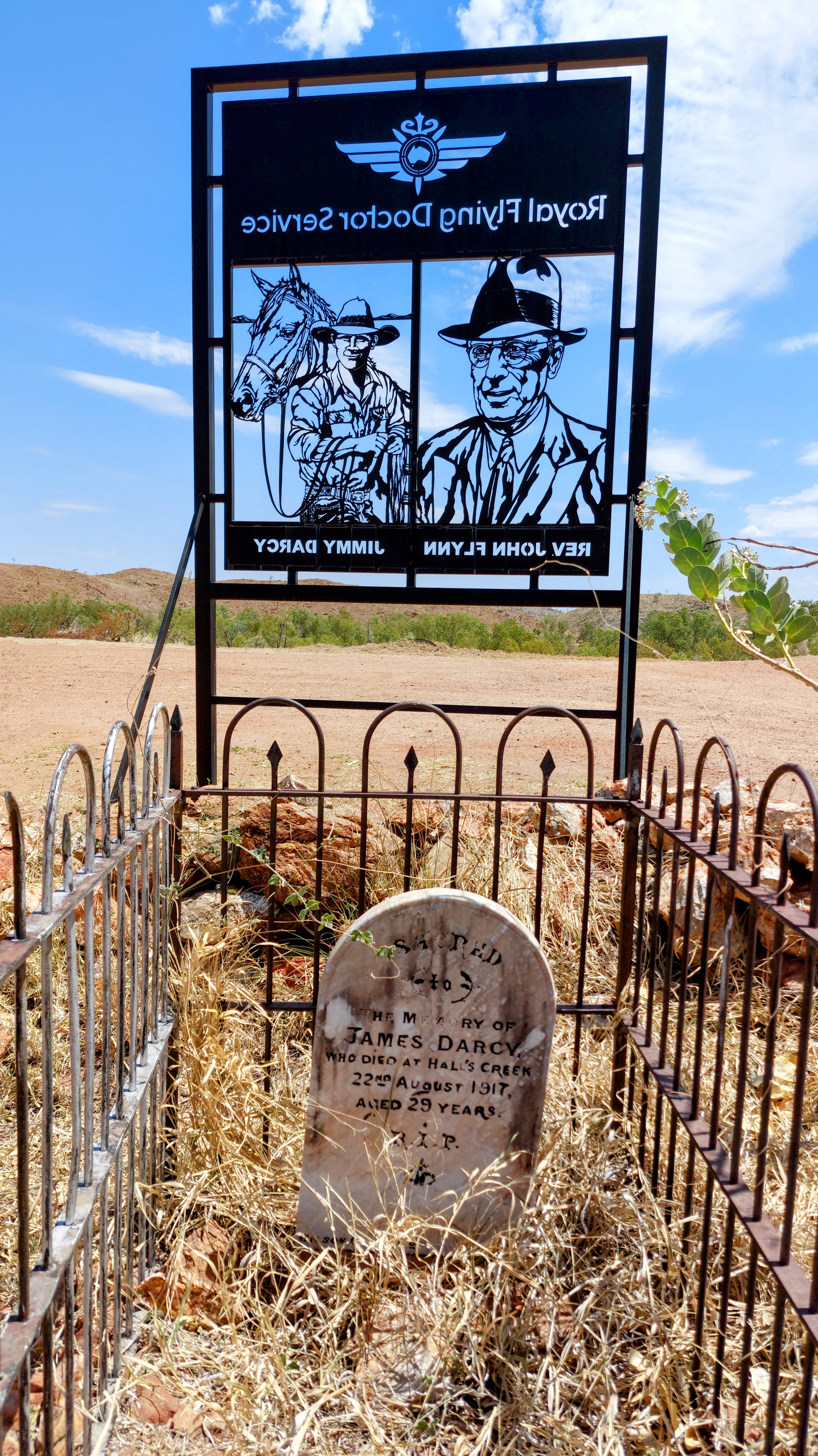 Jimmy Darcy's grave surrounded by a small fence. A large cutout metal sign highlighting both Darcy and the Rev John Flynn stands outside the desolate and sparse graveyard