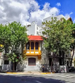 Casa en esquina con techo de tejas rojas,bordeada de árboles y con el cielo con nubes