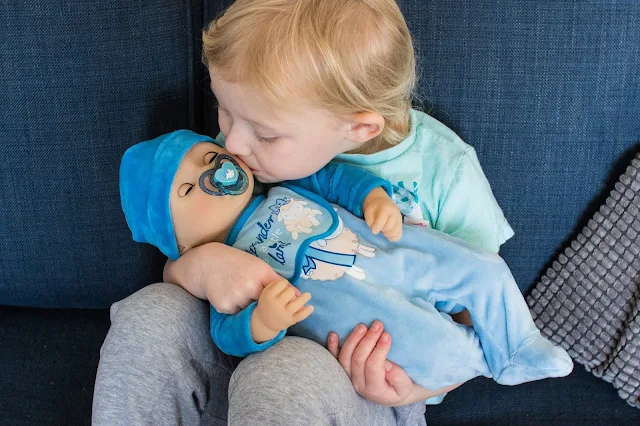 A 4 year old girl sitting on a sofa holding a Baby Alexander doll with a dummy in, she is kissing the doll on it's cheek