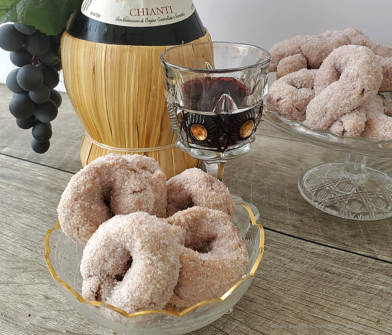 these are an round Italian cookie made with red wine and coated in sugar in a glass dish with a bottle of chianti and more cookies i n the background