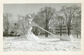 A black and white photograph showing a tall snow sculpture of a short man blowing on an oversized horn.