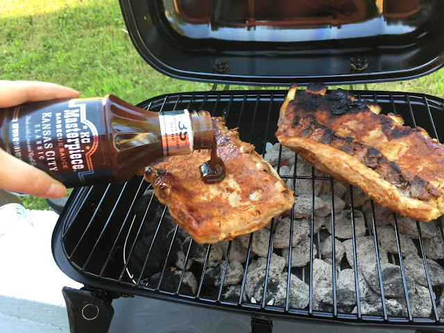 The ribs on the grill, with BBQ sauce being poured over the bottom of the half rack.