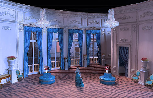 Photo of a set model. The model is of a spacious room whose back wall faces a street and includes four floor-to-ceiling windows with blue drapes that sweep the floor. There are a few turquoise chairs against the walls and two turquoise circular couches. There's a door in a wall to the right and another to the back of the set.