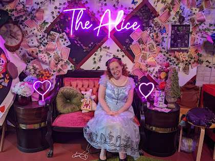 The Princess Blogger dressed as Alice sitting in the Alice bench under glowing neon sign for The Alice