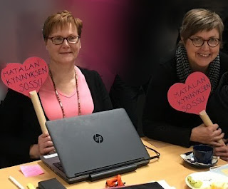 Seija Okulov ja Anne Tuikka istuvat pöydän ääressä käsissään "Matalan kynnyksen sossu" -kyltit.