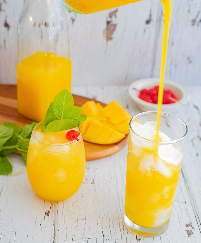 Pouring a glass of mango juice with another filled glass garnished with mint and a cherry.