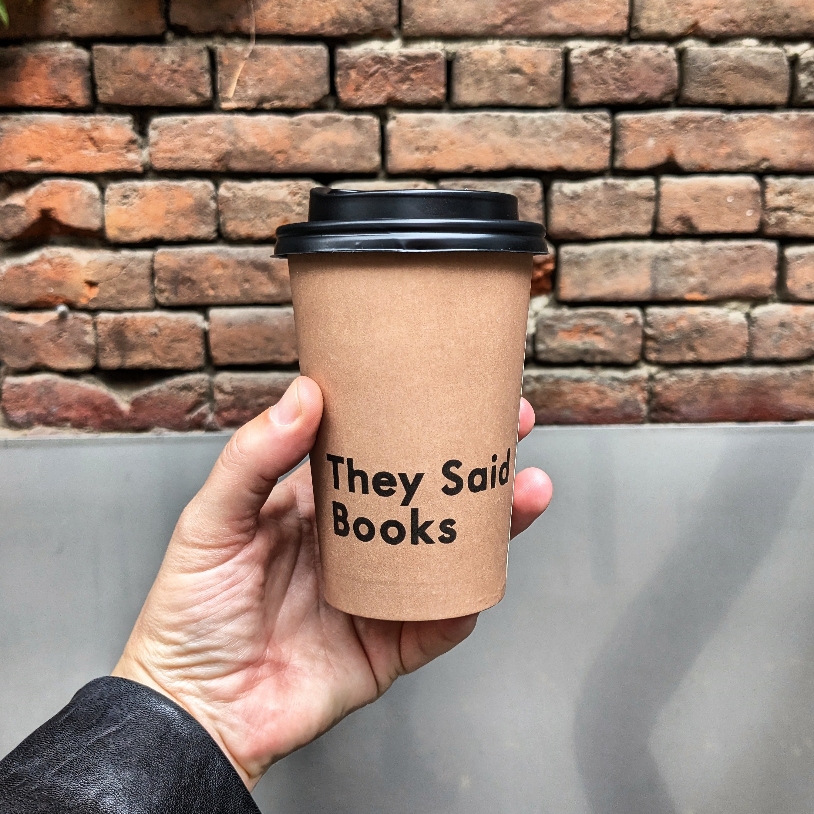 Take out coffee being held up against a brick wall. The coffee is from They Said Books, one of the best cafes in Tbilisi that sits above a book shop