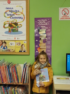 Dziewczynka stojąca przy ścianie na której wiszą materiały i plakaty promujące akcję Mała Książka -Wielki Człowiek. Dziewczynka trzyma w ręku pluszowa sowę i książkę.