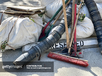 A pile of basic landscaping tools, including shovels, rakes, and pots in front of a reserved parking space..