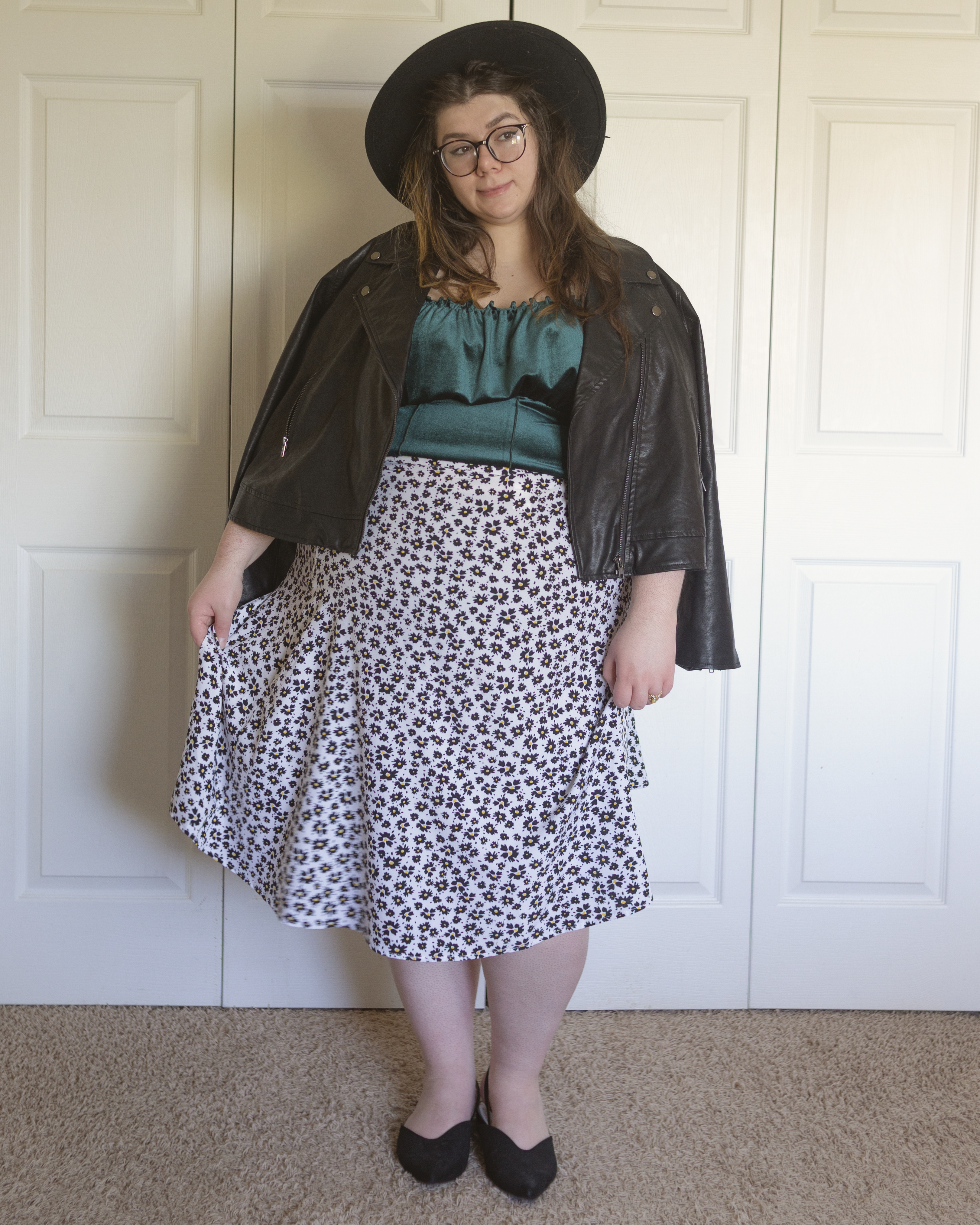 An outfit consisting of a black wide brim porkpie hat, a black faux leather moto jacket draped over the shoulders on top of a dark green velvet camisole top tucked into a black on white daisy print midi skirt and black slingback pointed toe flats.