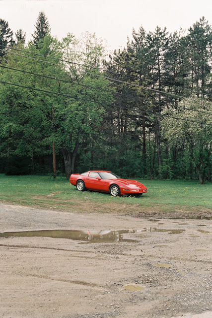 from the series 'Long Term Parking' by photographer Nicolas Poillot.  after the rain, a red corvette is parked on the lawn, grass, powerlines, springtime