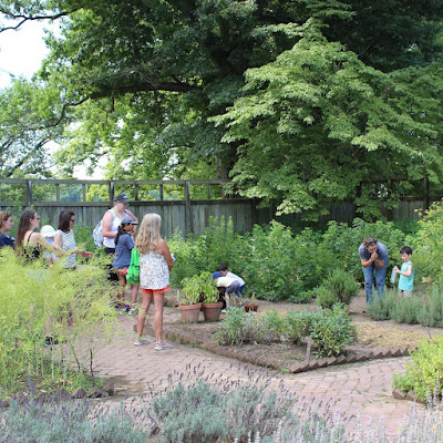 Children and adults stand on brick pathways that run between garden beds with herbs and flowers