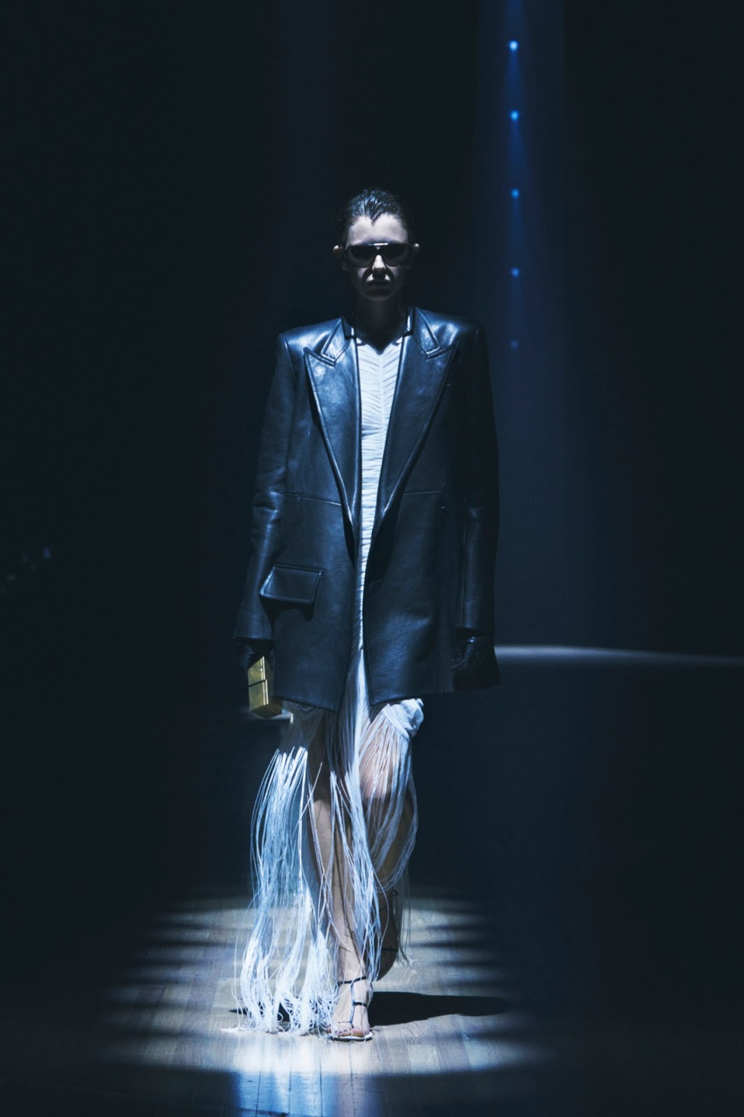 Woman with dark brown hair in sleek bun wearing aviator sunglasses, an oversized black leather blazer, and a beige maxidress with fringe carrying a gold metal clutch walking on wooden floorboards under a spotlight