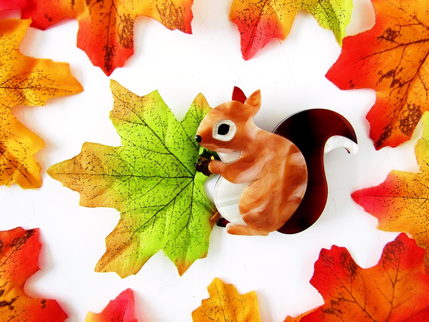 A photo of Erstwilder's Bright Eyed and Bushy Tailed red squirrel brooch, taken from above. The photo shows a reddish-brown squirrel brooch facing to the left and holding a little brown acorn in its paws. It sits on a white background surrounded by fiery faux autumn leaves.