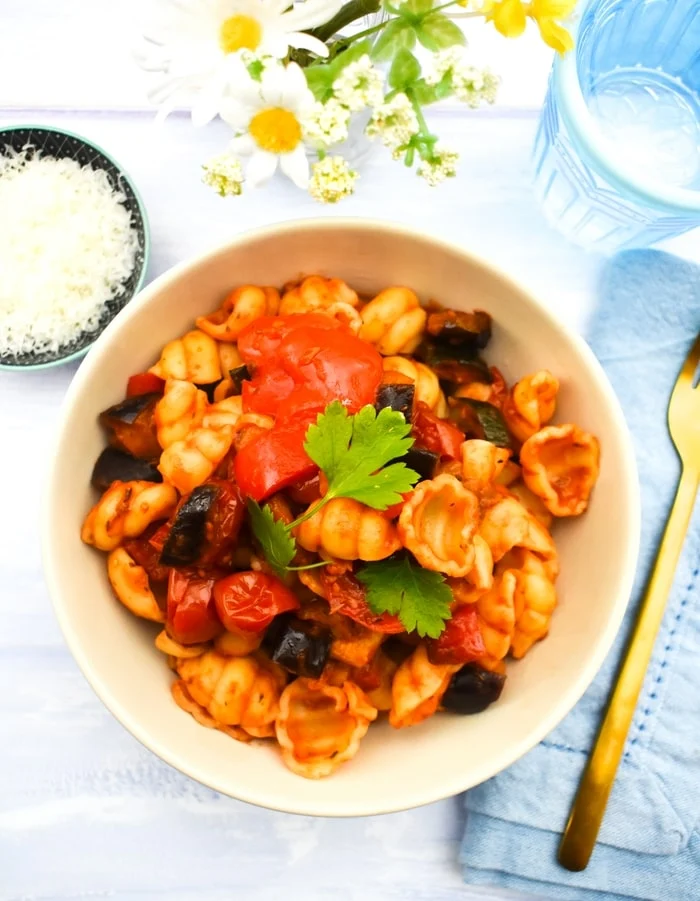 Ratatouille pasta in a pink pasta bowl next to yellow flowers and a glass of water