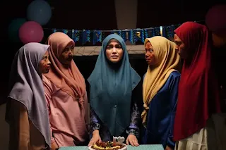 Seorang wanita berjilbab dikelilingi empat anak perempuan berwajah luka dalam suasana pesta ulang tahun yang menyeramkan.