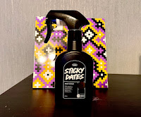 And why did an uncle photo of a big black spray bottle with a white dorm, engraved into the front of the bottle with sticky dates body, spraying white big font with Lush, in small white font, with an oval around it on a background of a geometric multi placard on a large, rectangular,, black shelf on a bright background