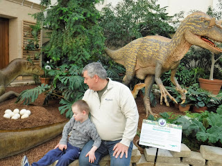 a man and boy sit together on a stone wall with dinosaur statues and plants behind them
