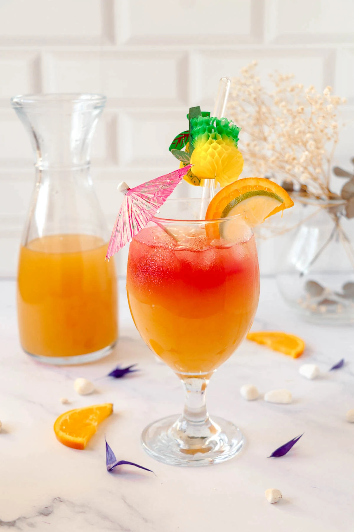 A glass filled with delicious Caribbean rum punch, garnished with umbrellas and citrus slices on a white background, with a bottle of rum punch in the background.