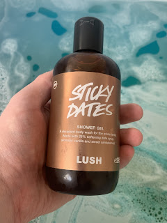 A white hand holding a clear cylindrical plastic bottle filled with dark brown liquid with a gold rectangular label with sticky dates shower gel lush in white font in front of an azure blue and gold bath with a white rectangular bath tub on a bright background
