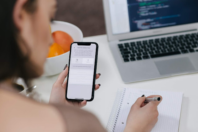 Mulher com celular na mão escrevendo em um caderno sobre a mesa em seu home office