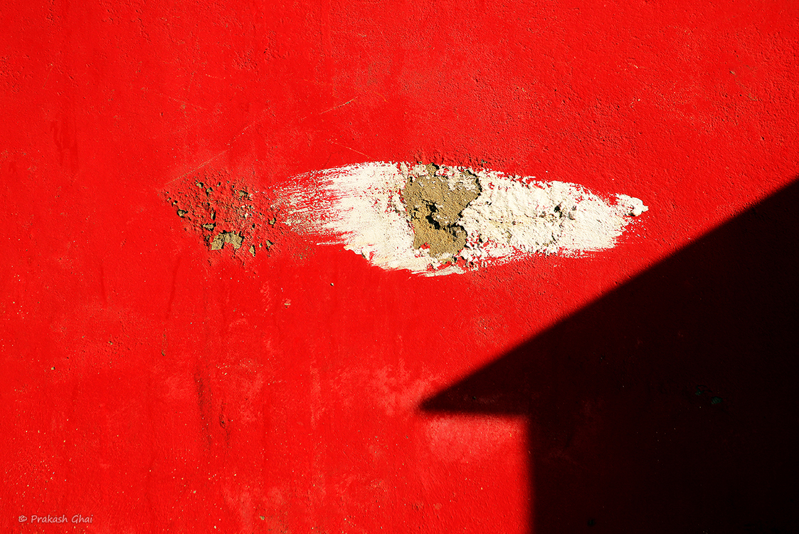 A minimalist photo of White scratch and shadow of a Hut shaped home on a red wall.