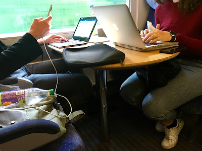 Zwei Menschen im Zug, jeweils am Laptop, einer hält ein Handy hoch