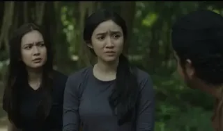 Tiga karakter dalam adegan film tengah berdiskusi di hutan, dua perempuan muda tampak cemas dan penasaran mendengarkan seorang pria yang berbicara serius di hadapan mereka.