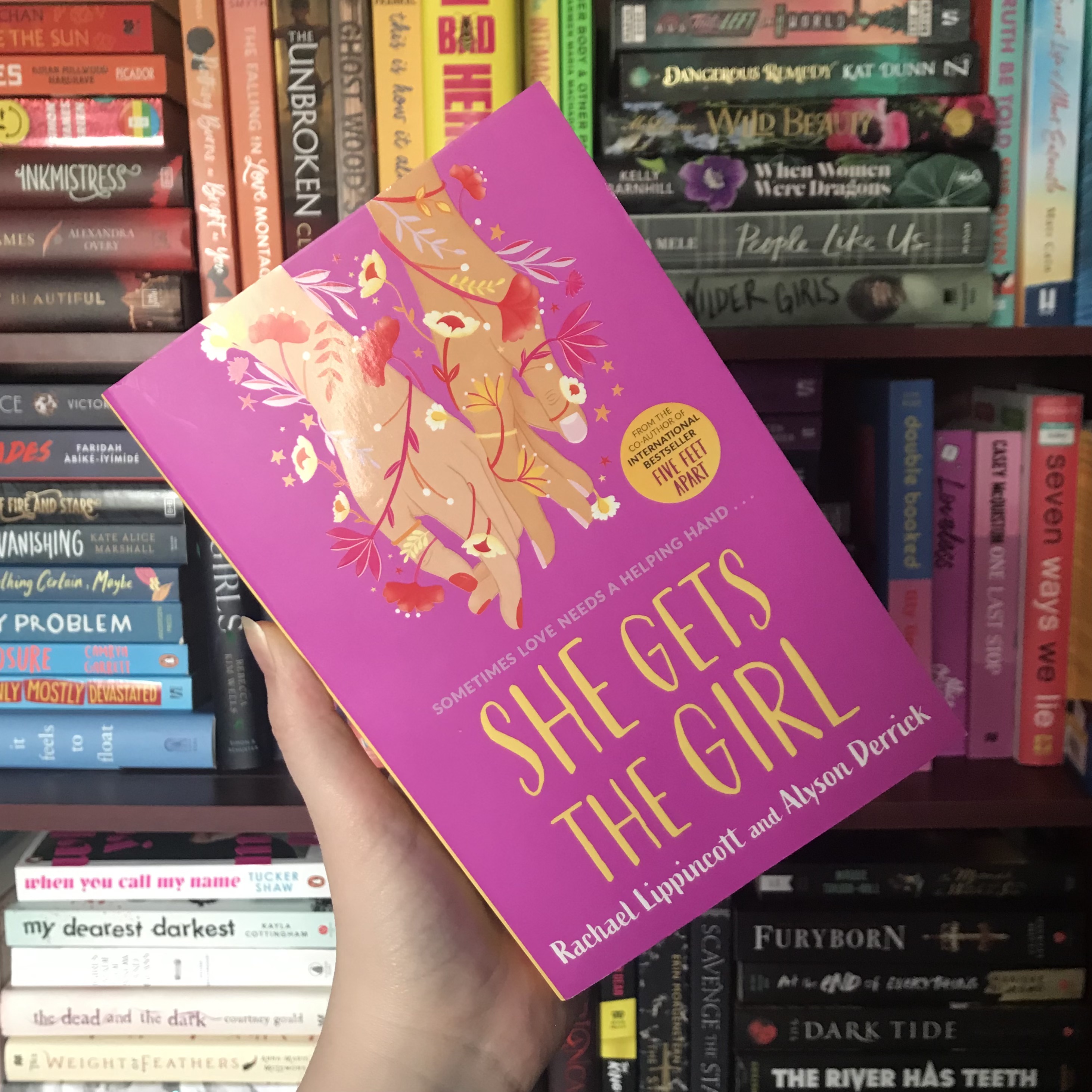 She Gets the Girl by Rachael Lippincott & Alyson Derrick is held up in front of rainbow shelves by a white hand.