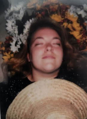 A young woman reclining on a pile of fake flowers, with her eyes closed and a faint smile, holding a straw hat over her chest.