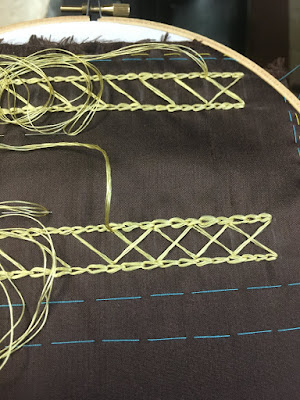 Dark brown silk stretched in a wooden embroidery hoop, with a pair of doubled gold chain stitch lines flanked by basted turquoise guidelines. The doubled lines are connected by diagonal gold stitches, and starting to be crossed into exes, with long coils of thread spooled on top of the work.