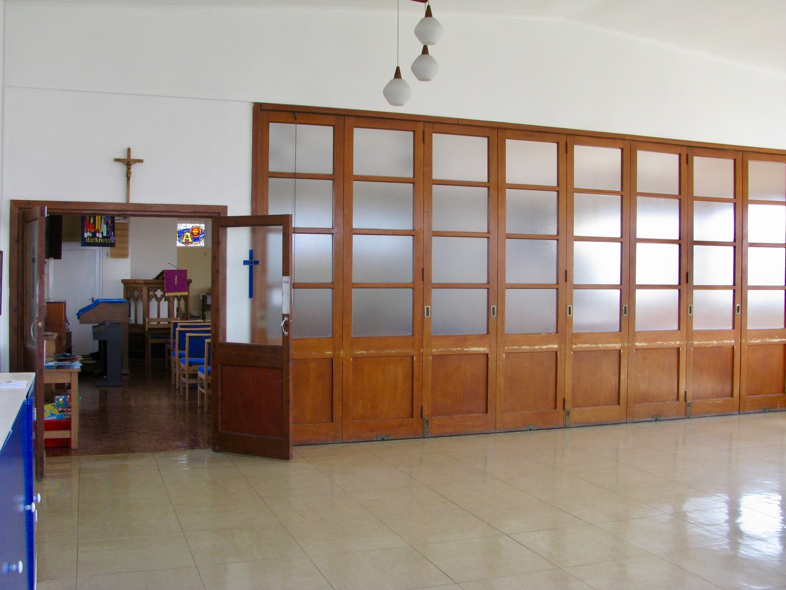 Image of entrance to church via wooden glazed doors and wooden glazed sliding doors which open to extend the church in to the hall. White walls. Sunny outside