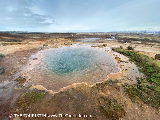 A bluish, green geothermal spring surrounded by brown-read mud and green moss.