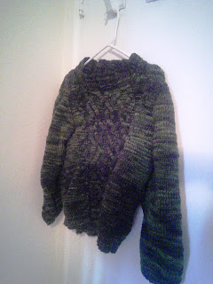 A dark green sweater hanging on a clothes hanger.  The front of the sweater has an aran cable pattern.