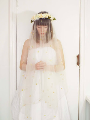 Ellie looking down, wearing a long white dress out of a bed sheet, with net curtain veil and flower crown.
