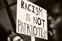 Black and white photograph showing a hand-drawn sign that reads "Racism is not Patriotism".